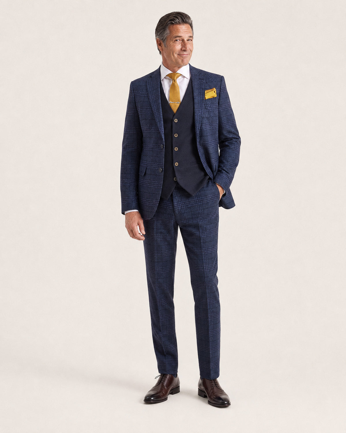 A man modeling a navy blue three-piece suit with a subtle windowpane check pattern, paired with a white dress shirt, a yellow tie, a yellow pocket square, and dark brown dress shoes, against a plain off-white studio background.