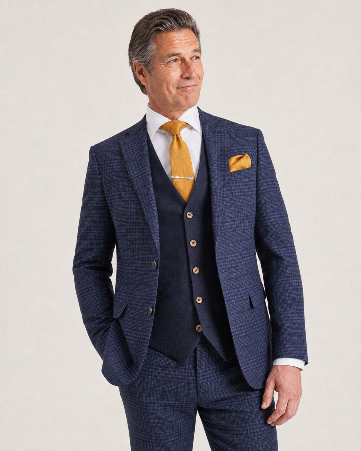 A man wearing a navy blue three-piece suit with a subtle windowpane check pattern, a matching navy vest, a white dress shirt, a gold tie, and a gold pocket square, standing against a light beige studio background.