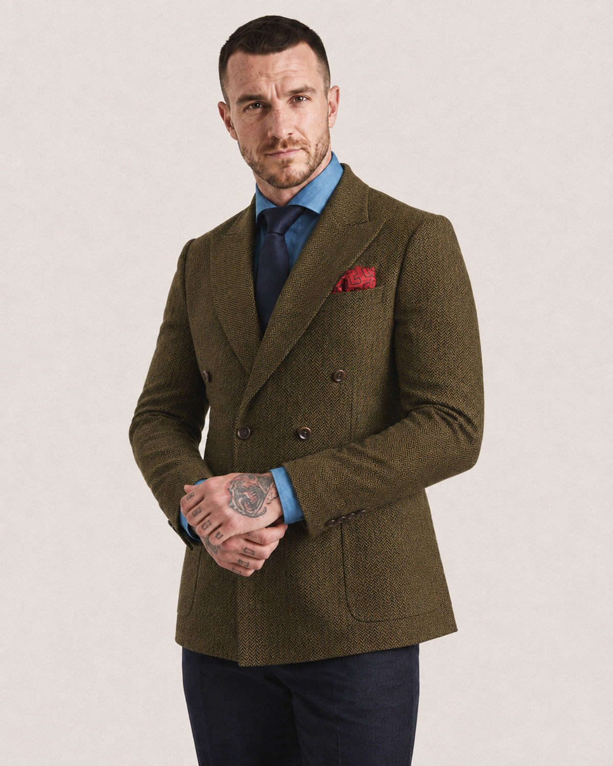 A man wearing a tailored double-breasted blazer in textured olive-brown tweed, styled with a light blue dress shirt, dark navy tie, and a red pocket square, standing against a plain light beige studio background.
