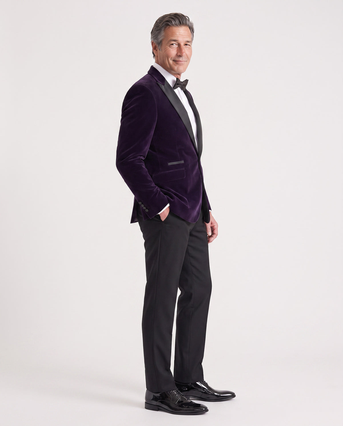 A man modeling a dark purple velvet tuxedo jacket with black satin lapels, paired with black trousers, a white dress shirt, a black bow tie, and black dress shoes, against a light gray studio background.