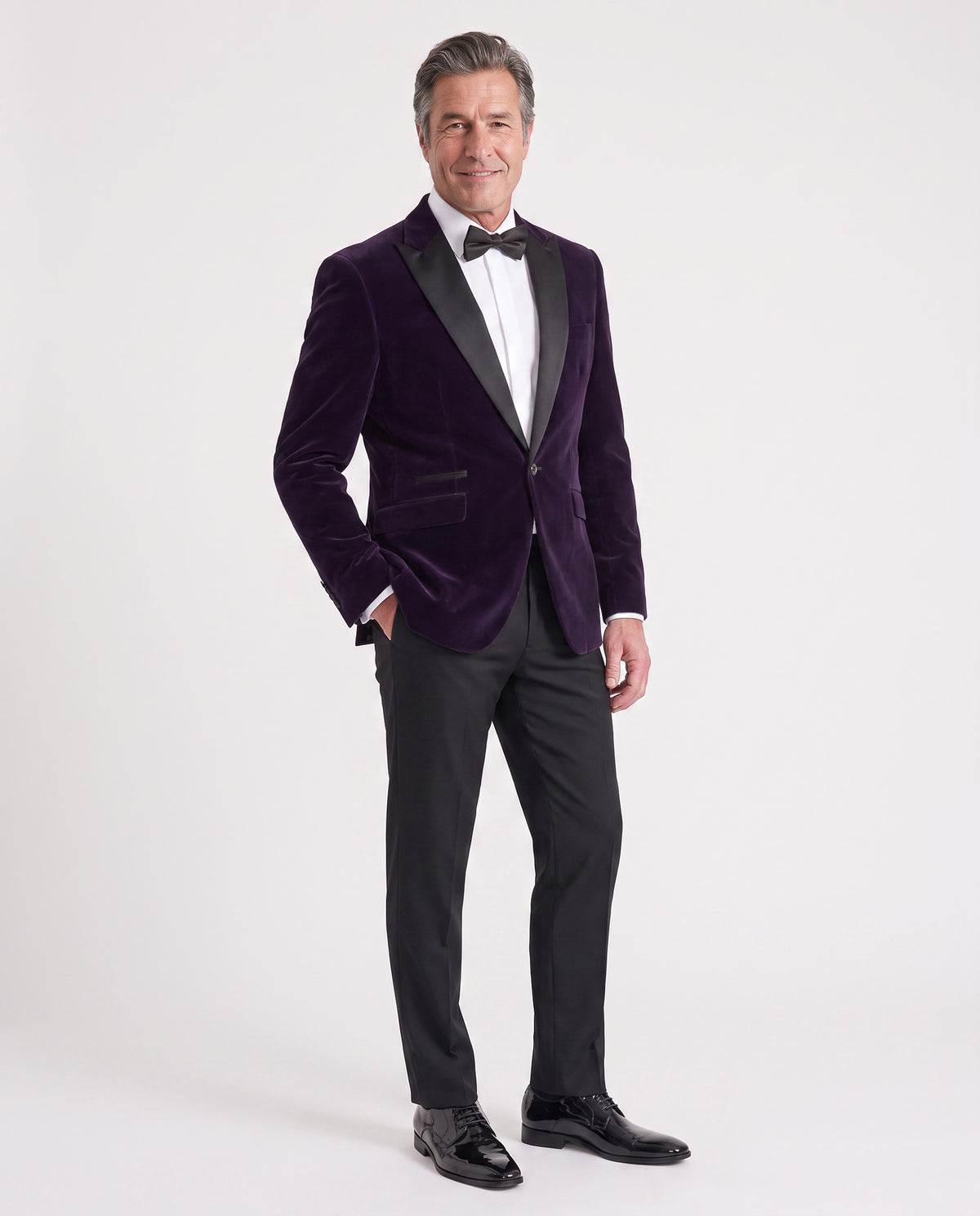 A man modeling a dark purple velvet tuxedo jacket with black satin lapels, paired with black trousers, a white dress shirt, and a black bow tie, against a light gray studio background.