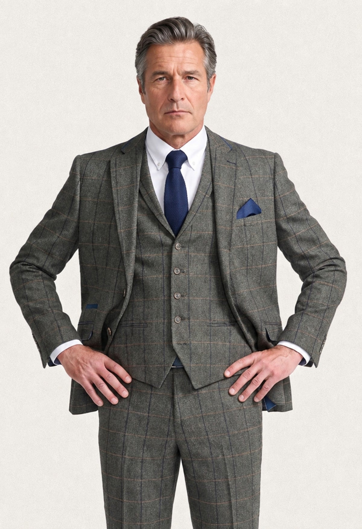 A man modeling a three-piece grey windowpane plaid suit with a white dress shirt, navy blue tie, and matching navy pocket square, set against a plain light background.