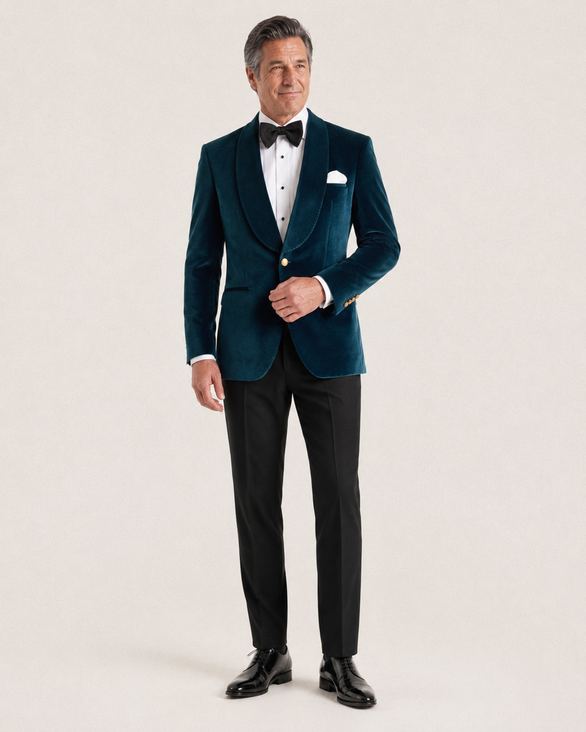 A man modeling a dark teal velvet tuxedo jacket with black shawl lapels, paired with black trousers, a white dress shirt, a black bow tie, and a white pocket square, against a light beige studio background.