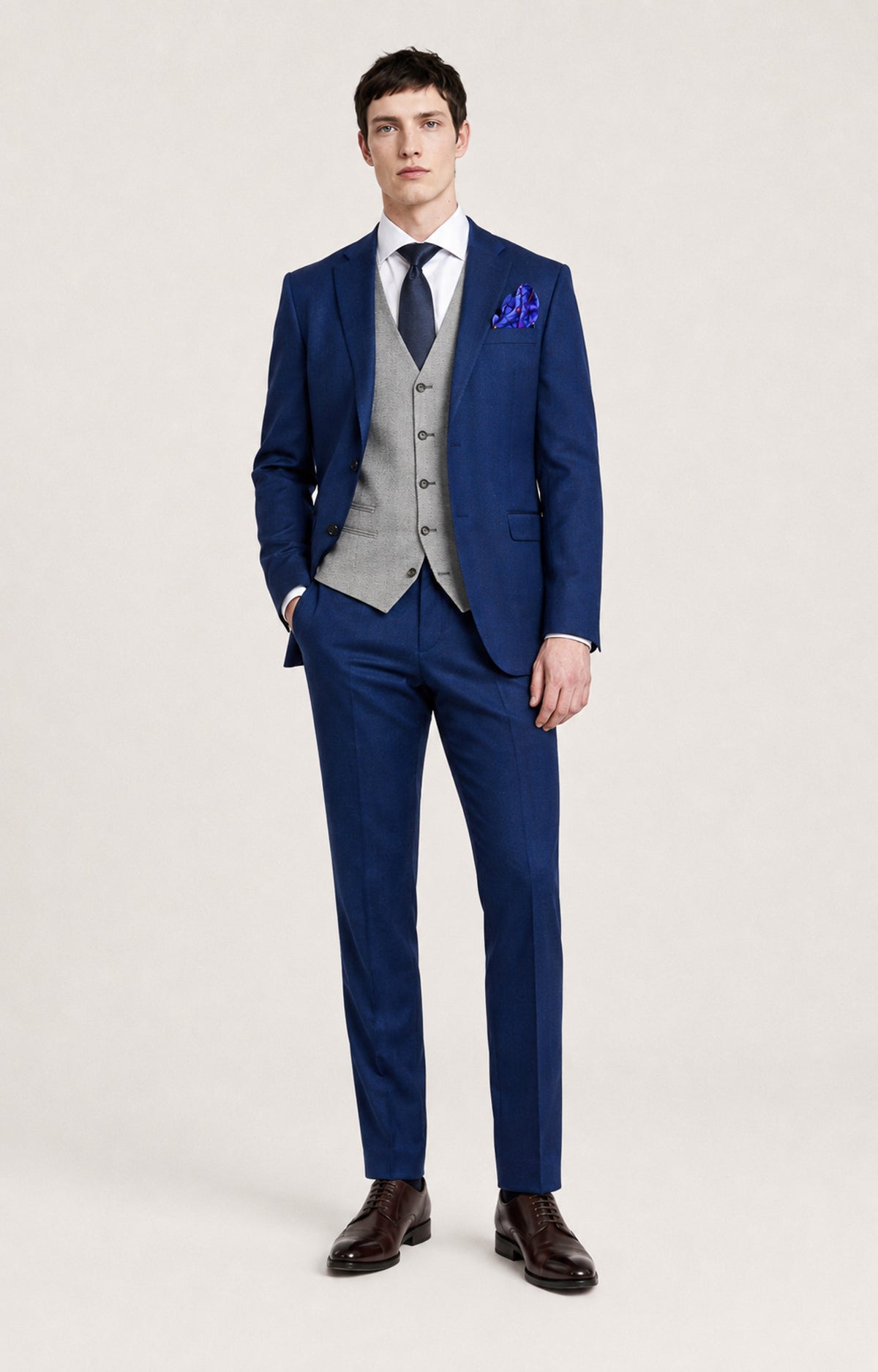 A man modeling a three-piece navy blue suit with a light gray patterned vest, a white dress shirt, a navy tie, and a blue patterned pocket square, standing against a light gray studio background.