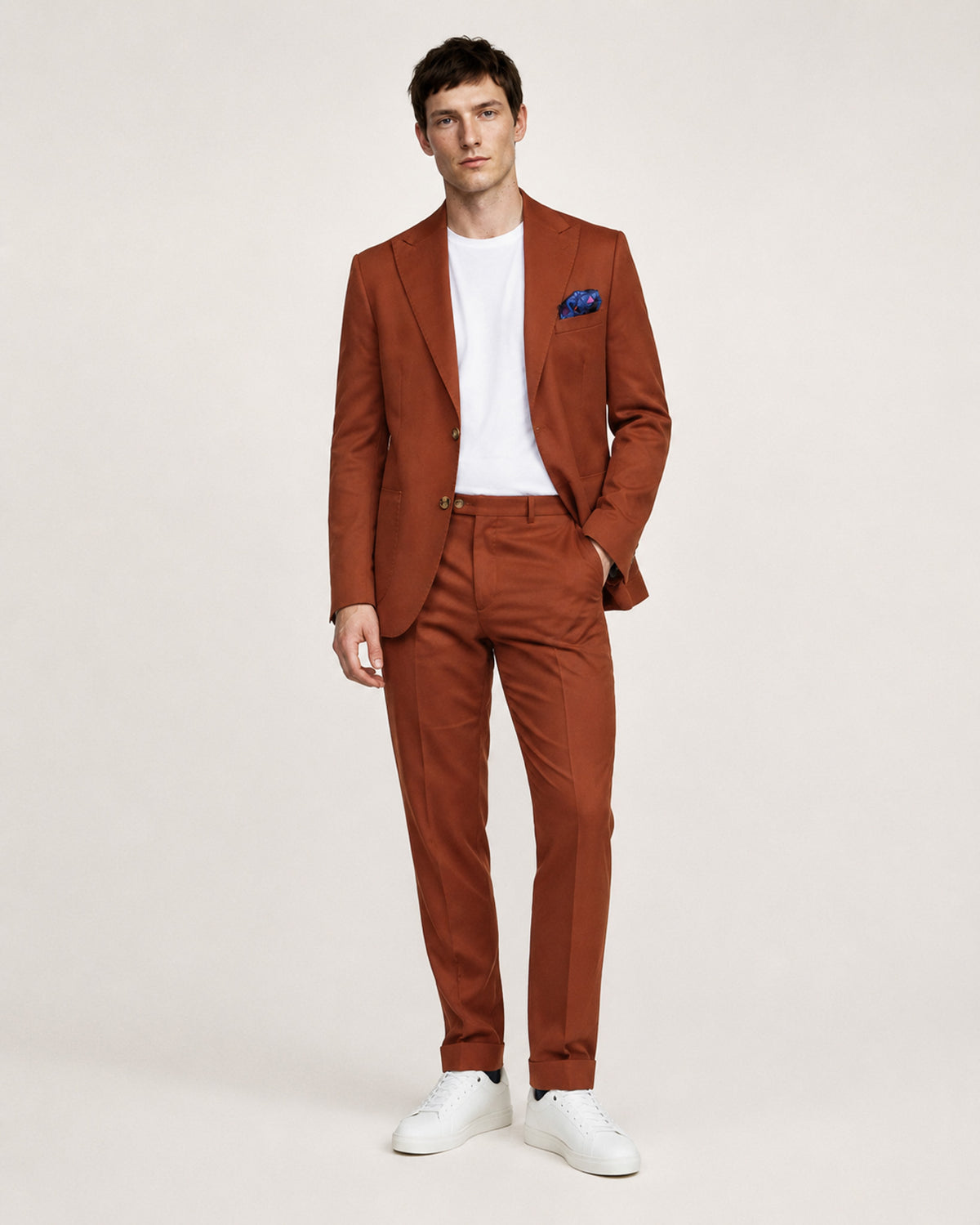 A man modeling a rust-orange tailored suit with a white crew-neck t-shirt, a blue patterned pocket square, and white sneakers, standing against a light beige studio background.