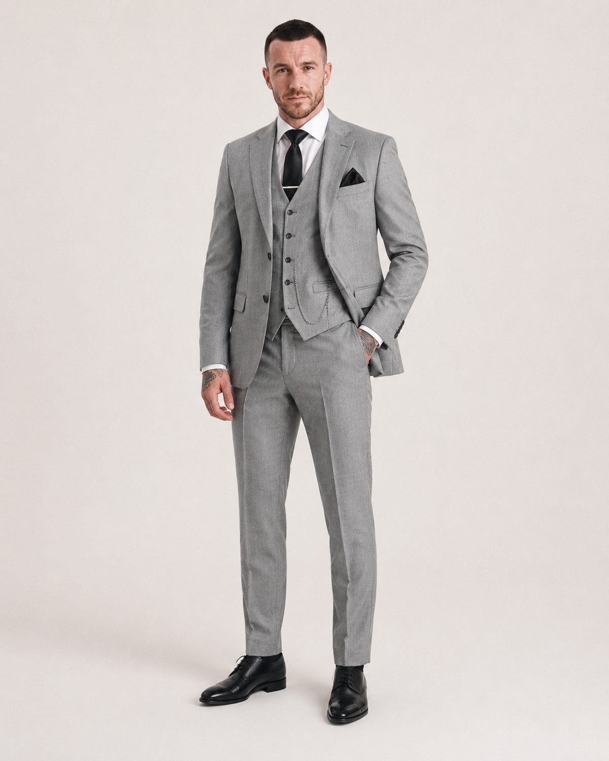A man modeling a light gray three-piece suit with a white dress shirt, black tie, and black dress shoes against a plain light beige studio background.