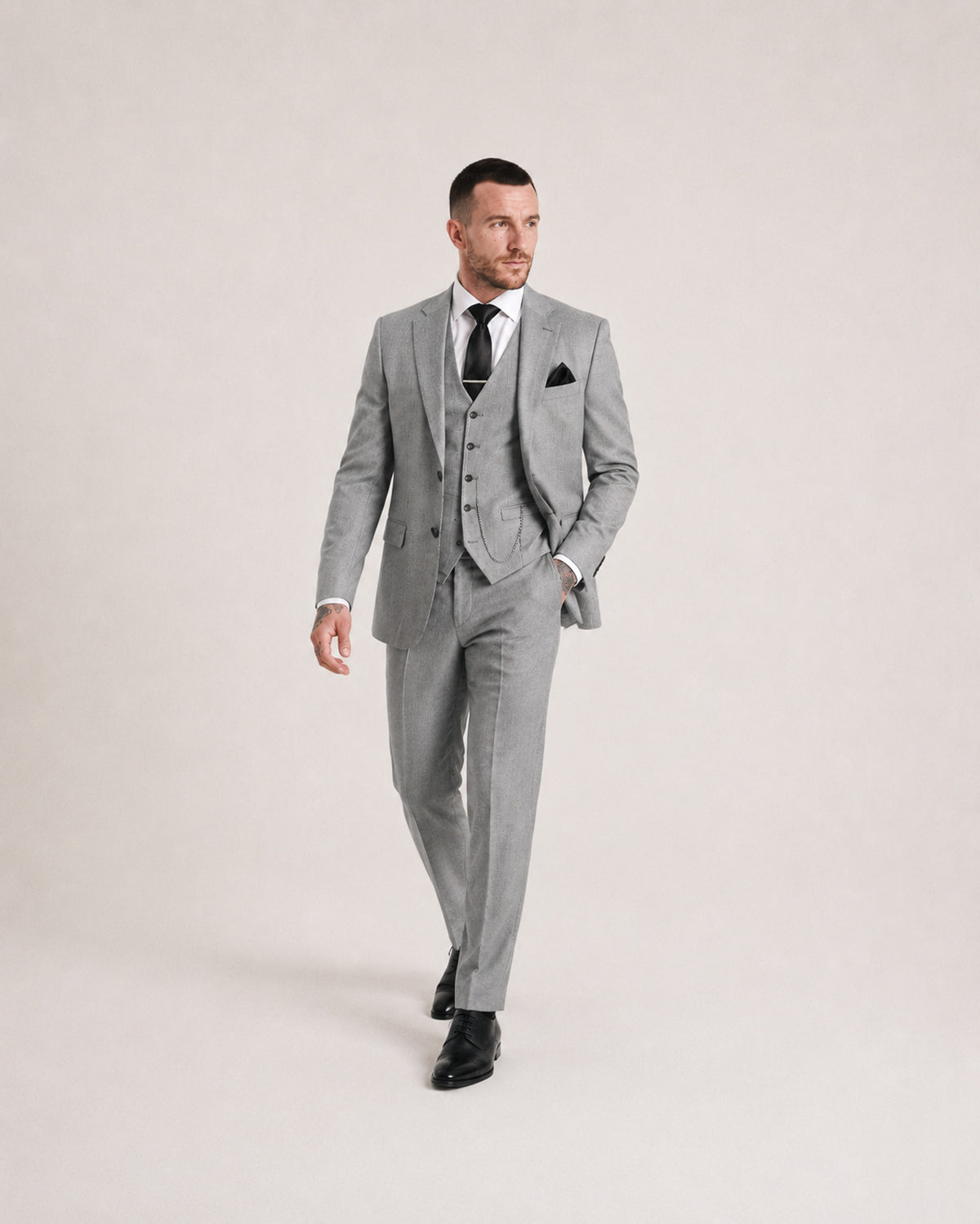 A man modeling a light gray three-piece suit with a matching vest and trousers, styled with a white dress shirt, black tie, and black dress shoes against a plain light beige studio background.