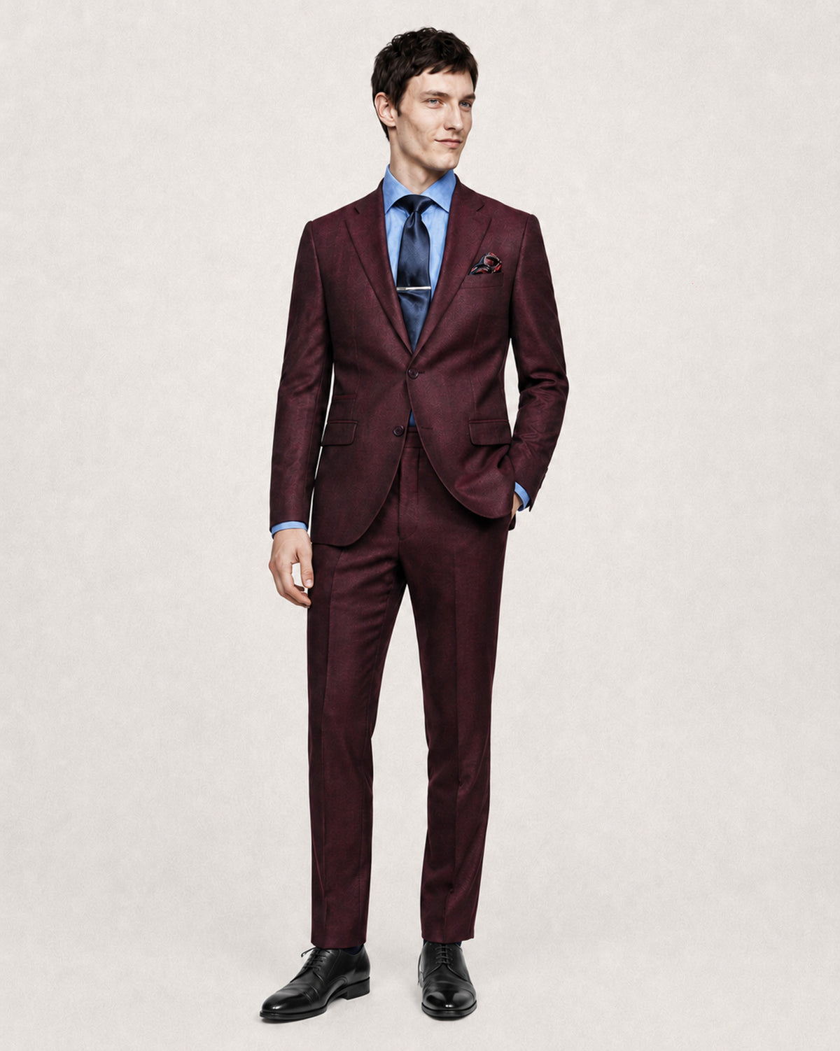 A man modeling a slim-fit two-piece suit in deep burgundy with a subtle micro-pattern, styled with a light blue dress shirt, a dark blue patterned tie, and black dress shoes against a light beige textured background.