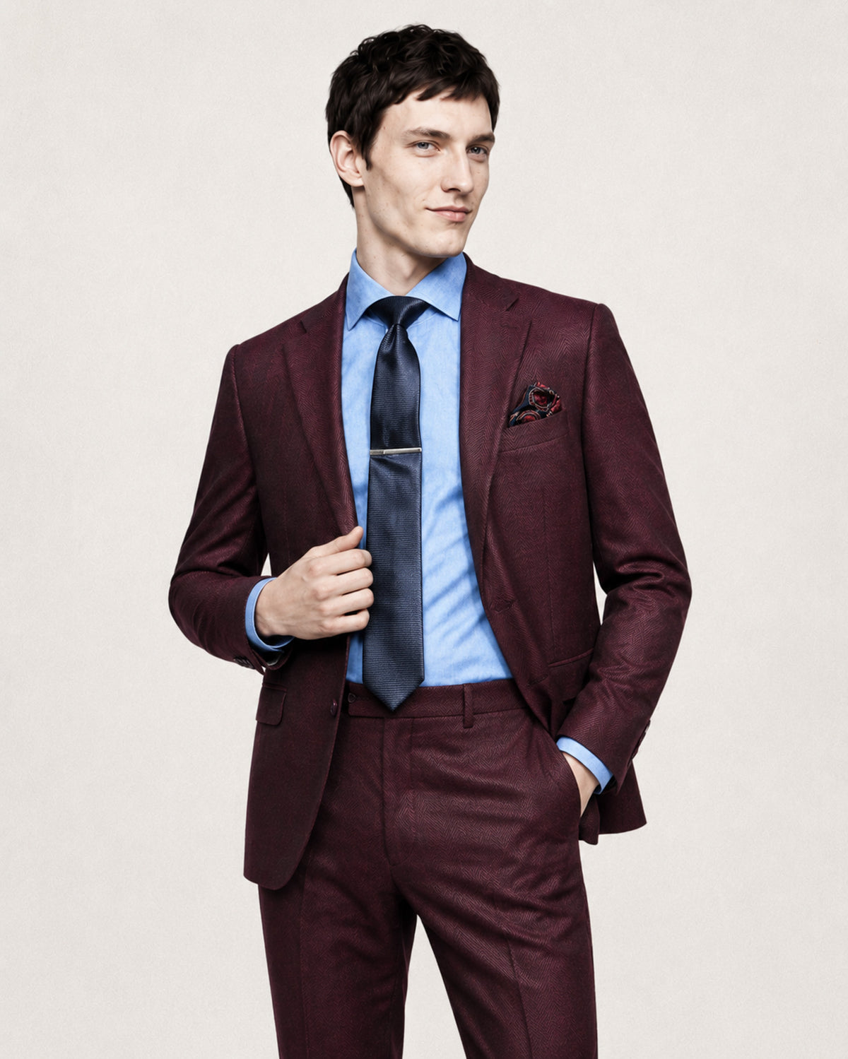A man wearing a burgundy suit with a light blue dress shirt, a dark blue tie, and a patterned pocket square, standing against a light beige background.