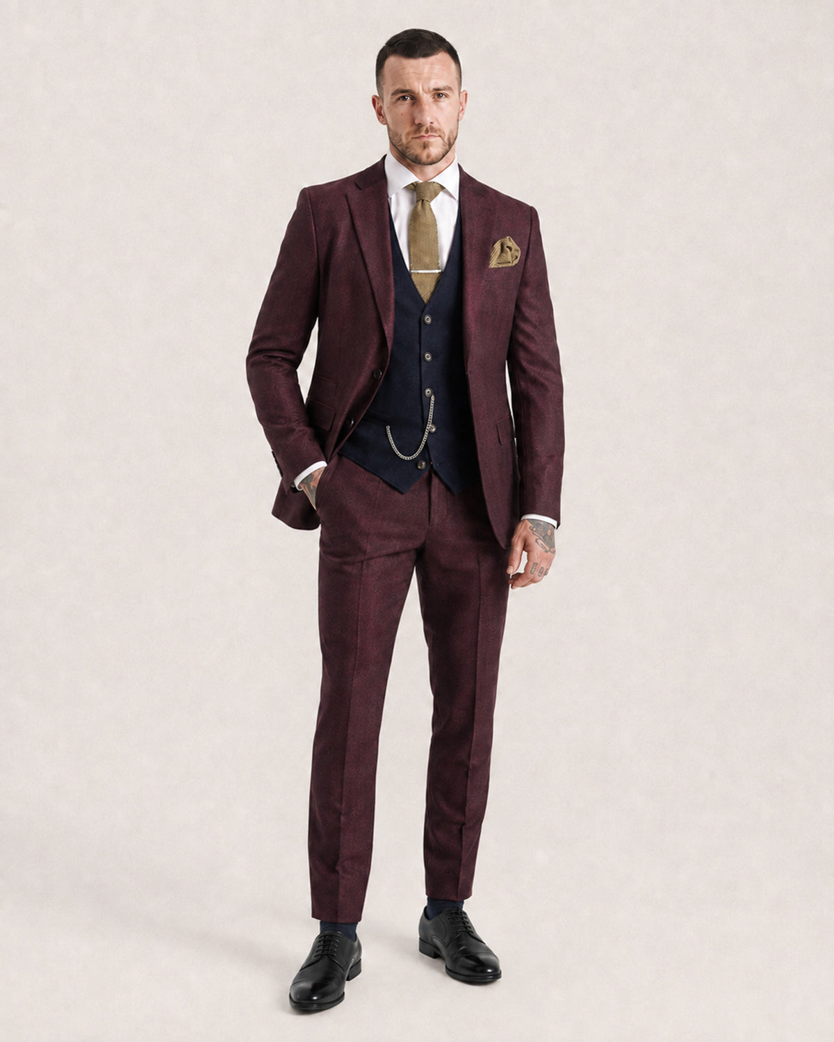 A man modeling a three-piece suit in a deep burgundy color, featuring a matching jacket, vest, and trousers, styled with a navy blue waistcoat, white dress shirt, gold tie, and a gold pocket square, set against a plain light beige background.