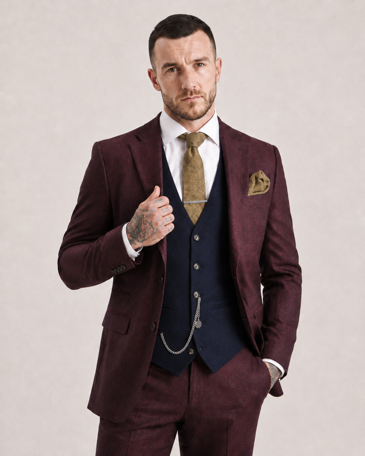 A man modeling a burgundy three-piece suit with a matching vest and trousers, styled with a navy blue waistcoat, a gold tie, a white dress shirt, and a matching pocket square against a neutral beige background.