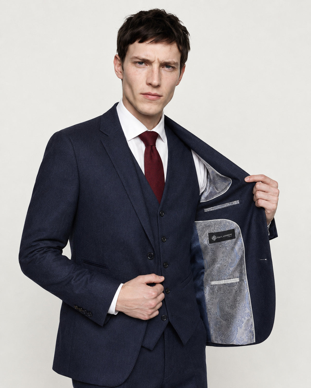 A man wearing a navy blue three-piece suit, white dress shirt, and burgundy tie, holding open his jacket to show the textured gray lining. Photographed against a clean, light off-white studio background.