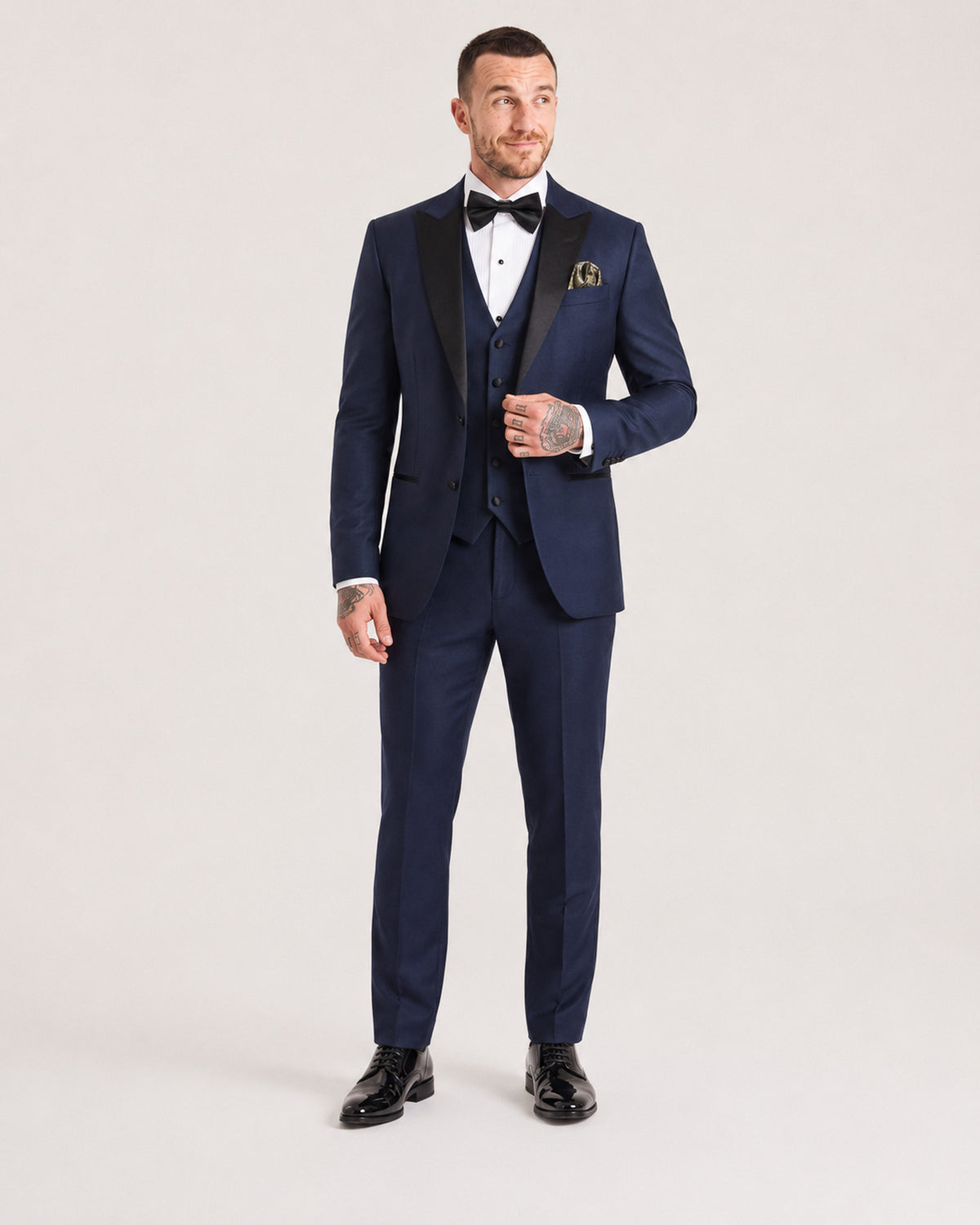 A man modeling a navy blue three-piece tuxedo with black satin lapels, a matching navy vest, white dress shirt, black bow tie, and a gold pocket square, standing against a light gray studio background.