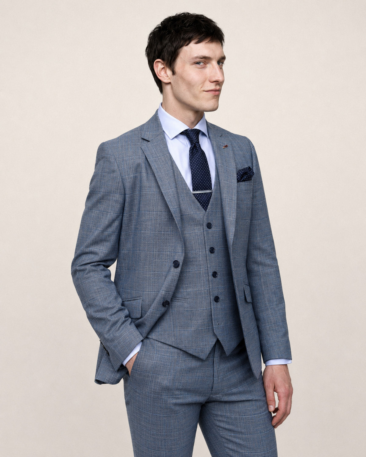 A man wearing a blue-gray three-piece suit with a subtle windowpane pattern, a light blue dress shirt, a dark navy patterned tie, and a matching pocket square, posed against a solid beige background.