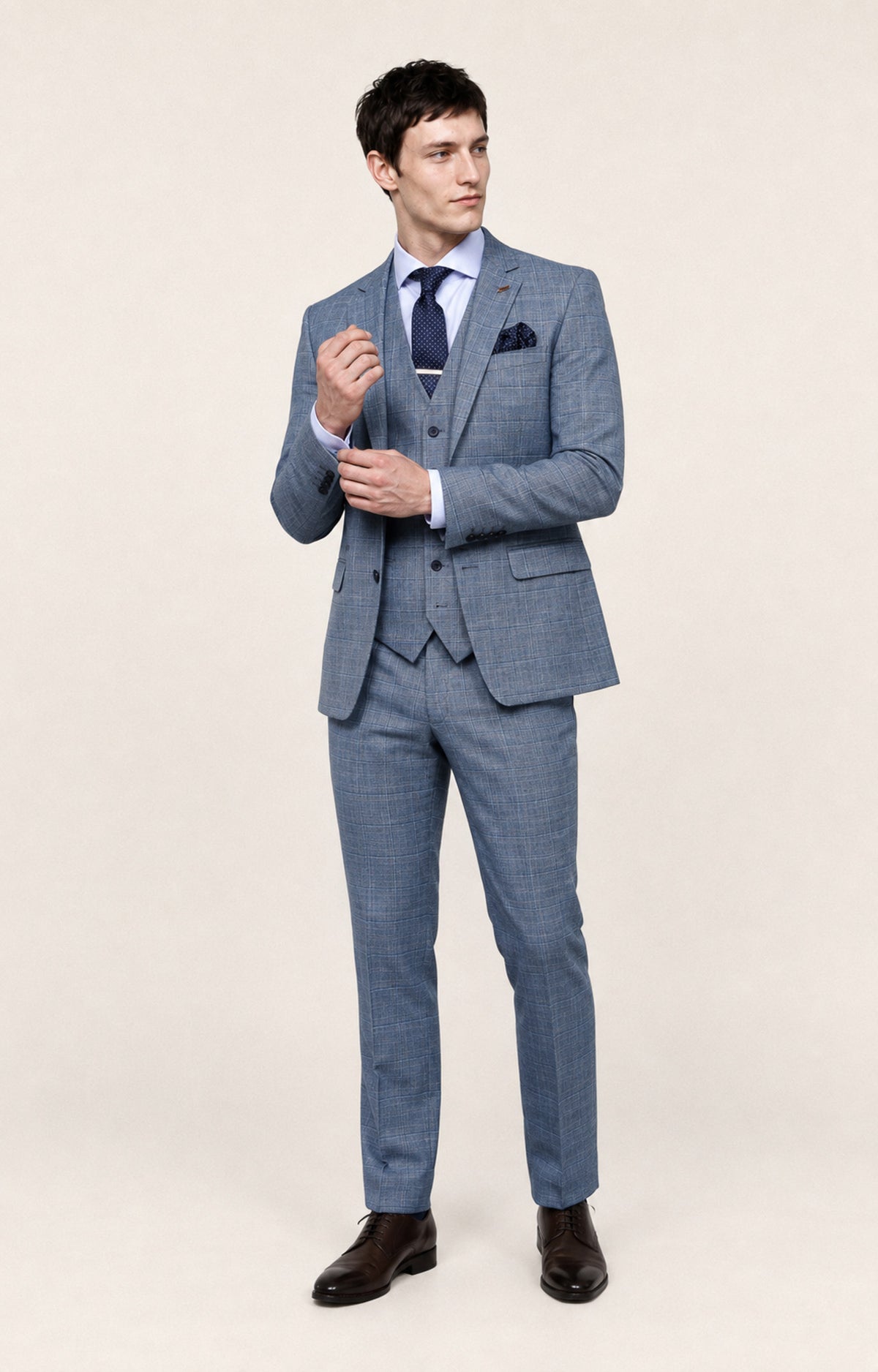 A man modeling a light blue three-piece suit with a subtle windowpane pattern, paired with a white dress shirt, navy tie, and dark brown dress shoes, against a solid beige studio background.
