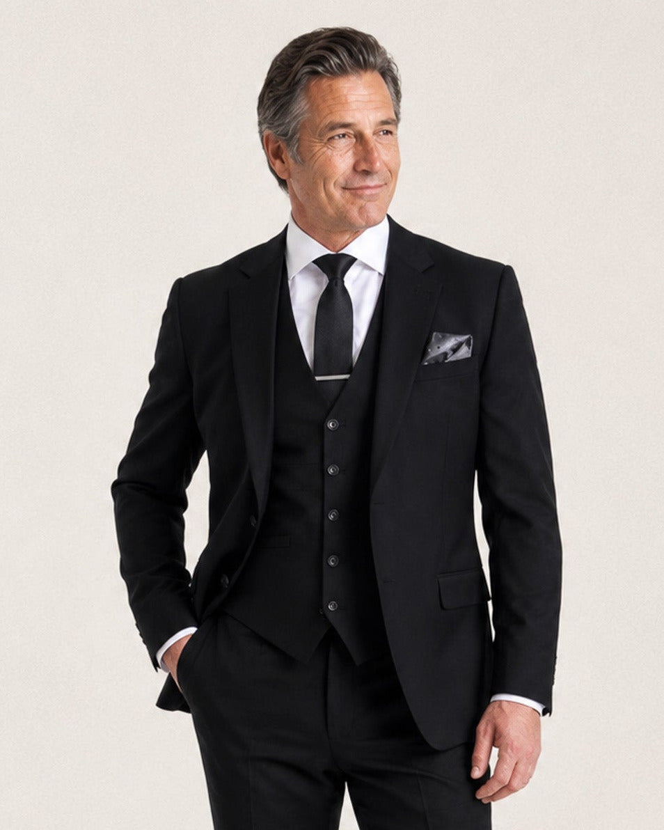A man modeling a black three-piece suit with a white dress shirt, black tie, and a patterned pocket square, standing against a light beige studio background.