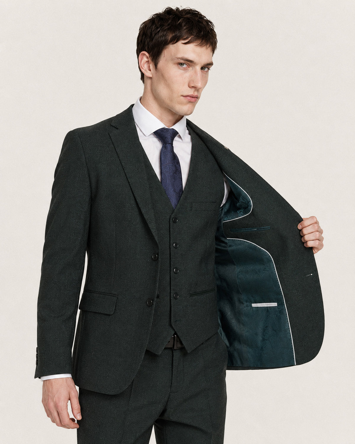 A man wearing a dark charcoal gray three-piece suit, holding open the jacket to show the dark teal lining. He is wearing a white dress shirt and a navy blue tie, photographed against a plain light beige background.