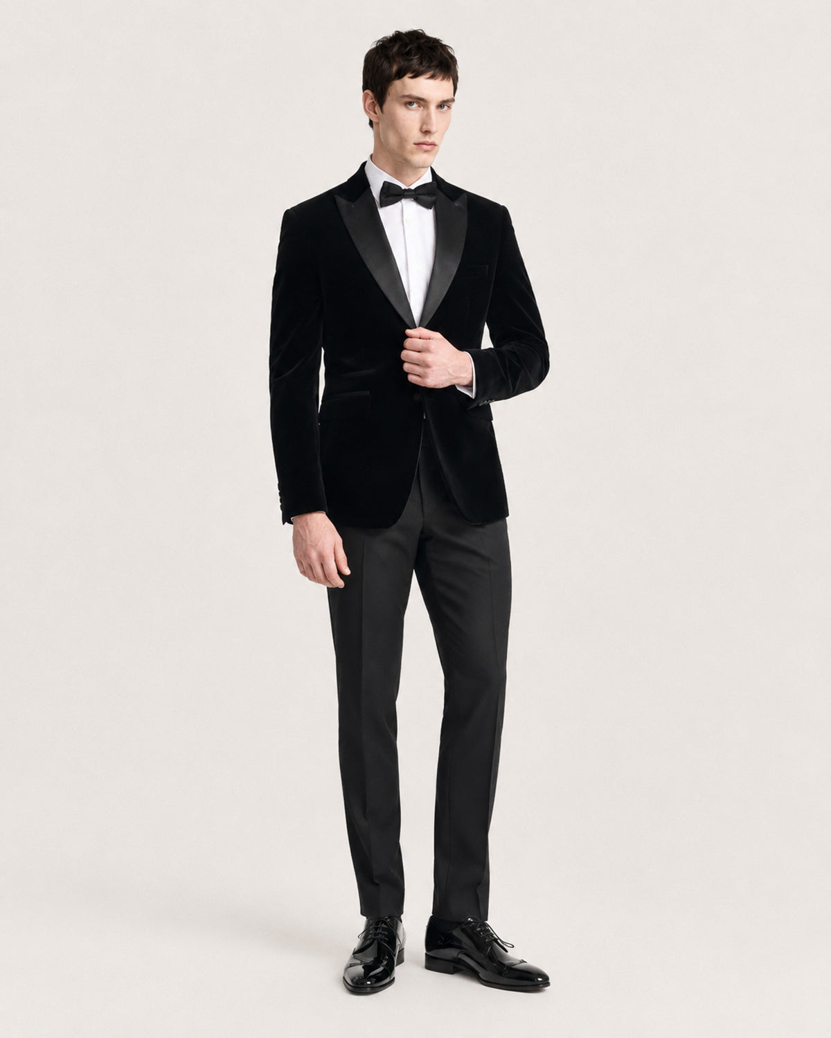 A man modeling a black velvet tuxedo jacket with black satin lapels, paired with black trousers, a white dress shirt, and a black bow tie, against a light gray studio background.