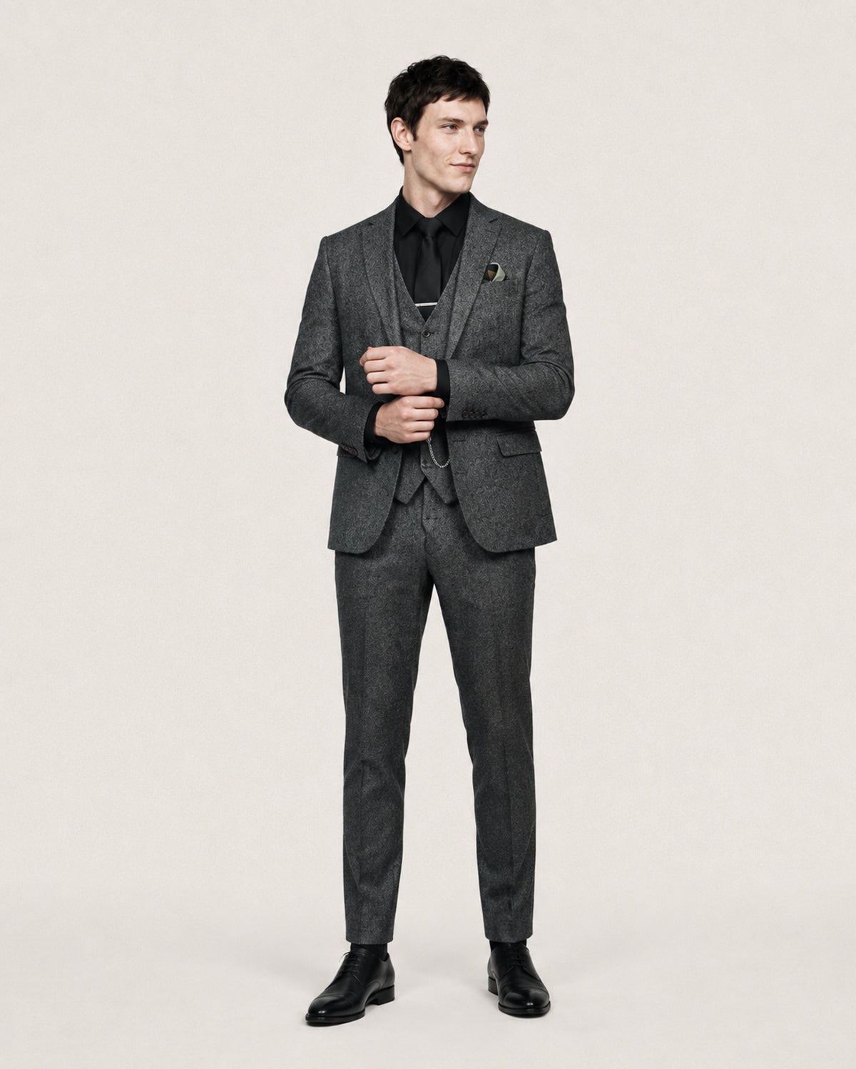 A man modeling a dark gray textured three-piece suit with a black shirt and black dress shoes, standing against a light beige studio background.