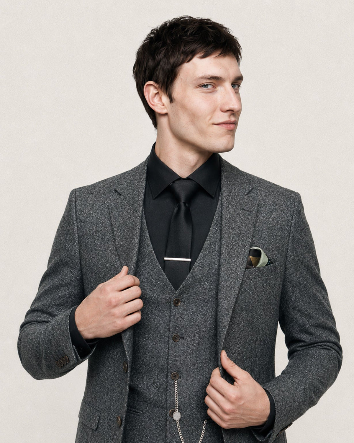 A man wearing a gray three-piece tweed suit with a black shirt, black tie, and black vest, standing against a light beige background.