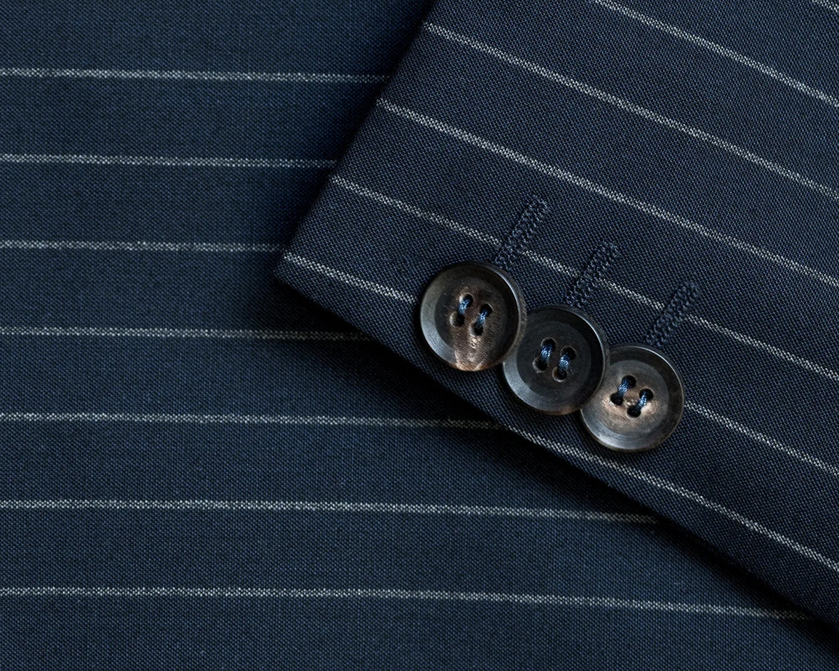 Close-up of a navy blue pinstripe fabric, likely a suit jacket or blazer, showing the texture of the weave, thin white vertical stripes, and three dark brown buttons on the cuff.