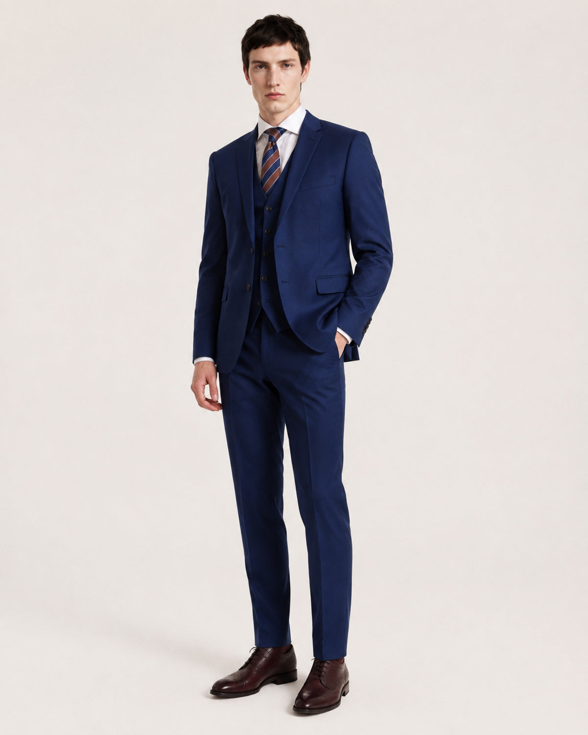 A man modeling a navy blue three-piece suit with a white dress shirt, a striped tie, and dark brown dress shoes, standing against a plain light beige studio background.