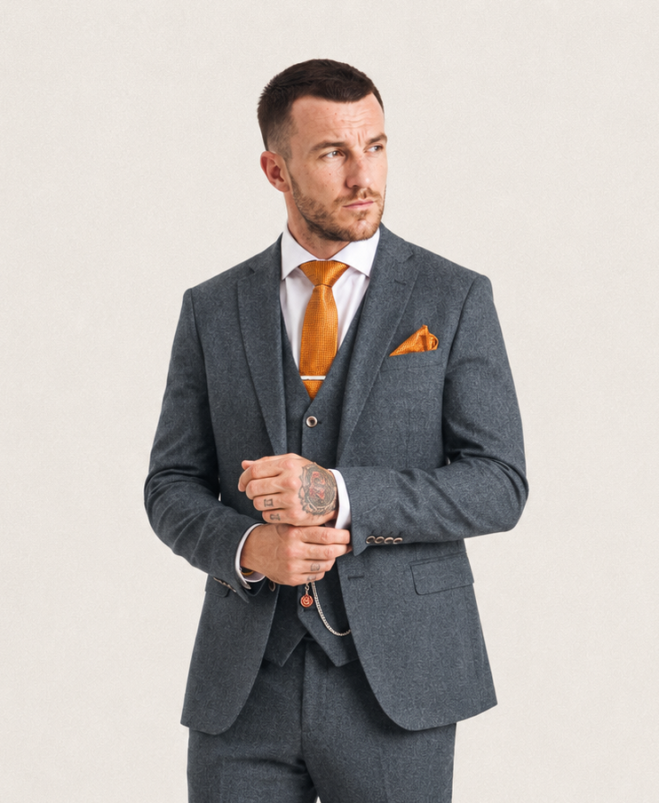 A man wearing a three-piece gray suit with a white dress shirt, orange tie, and orange pocket square, standing against a light gray background.