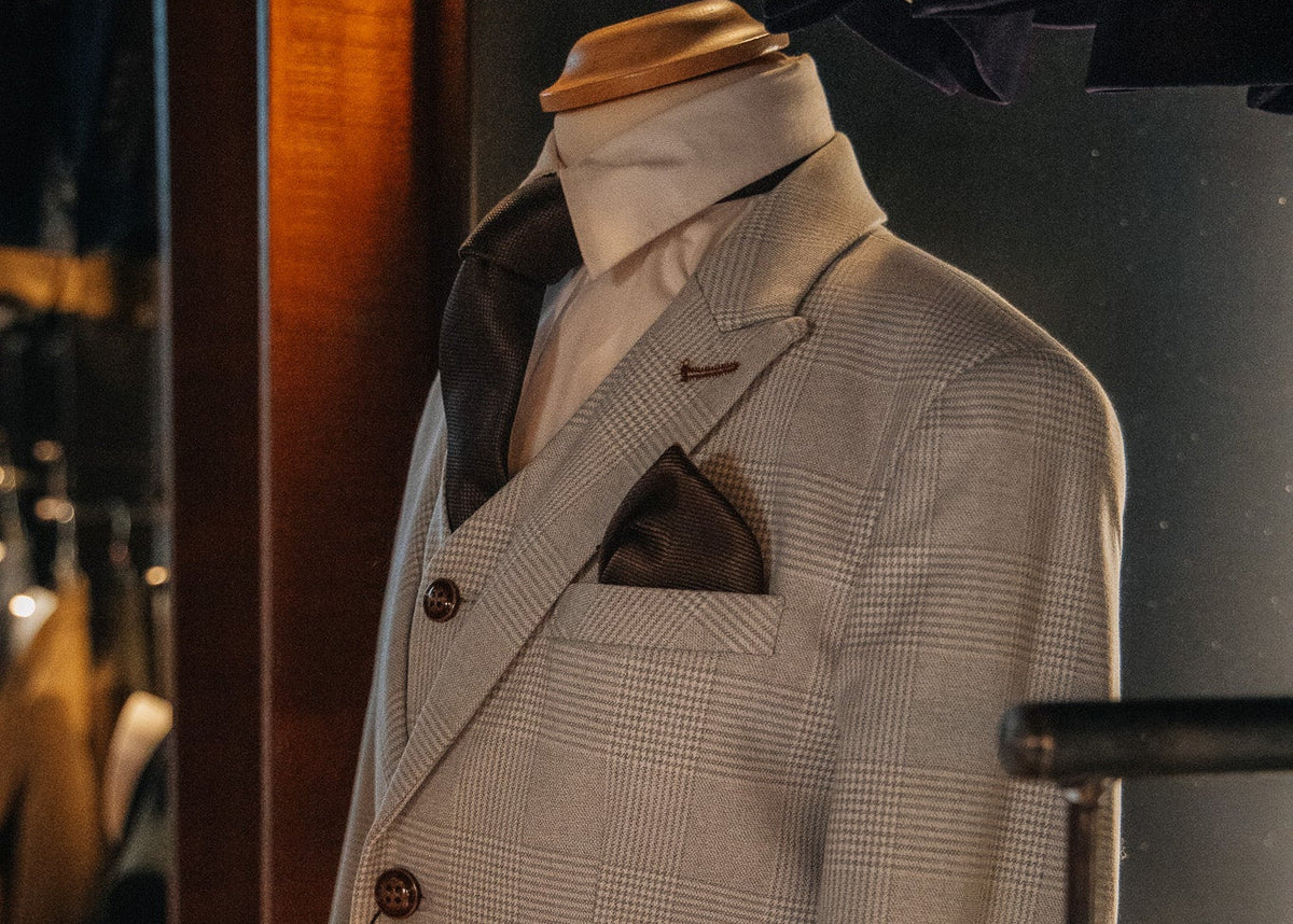 A light beige and white micro-check plaid suit displayed on a mannequin torso, featuring a three-button jacket, matching vest, trousers, a white dress shirt, and a dark brown pocket square.