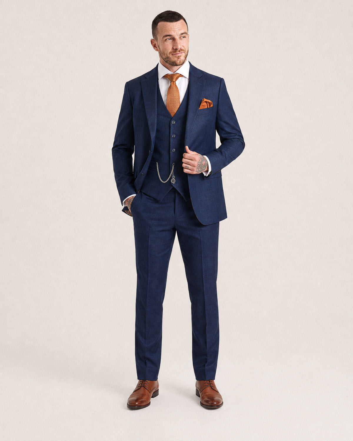 A man modeling a navy blue three-piece suit with a white dress shirt, orange tie, and orange pocket square, standing against a light beige studio background.
