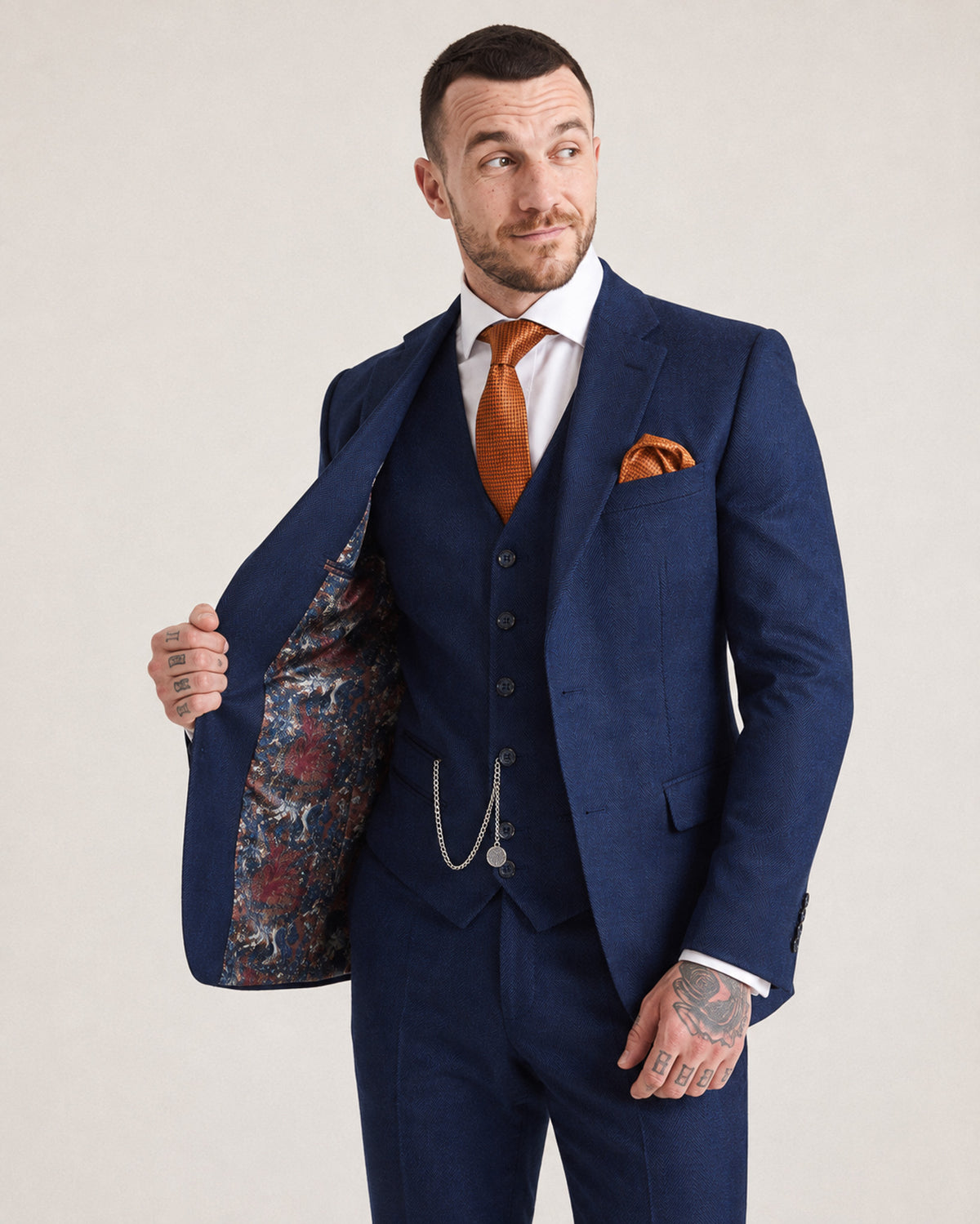 A man wearing a navy blue three-piece suit with a white dress shirt, orange tie, and matching orange pocket square, holding open his jacket to show the patterned lining.