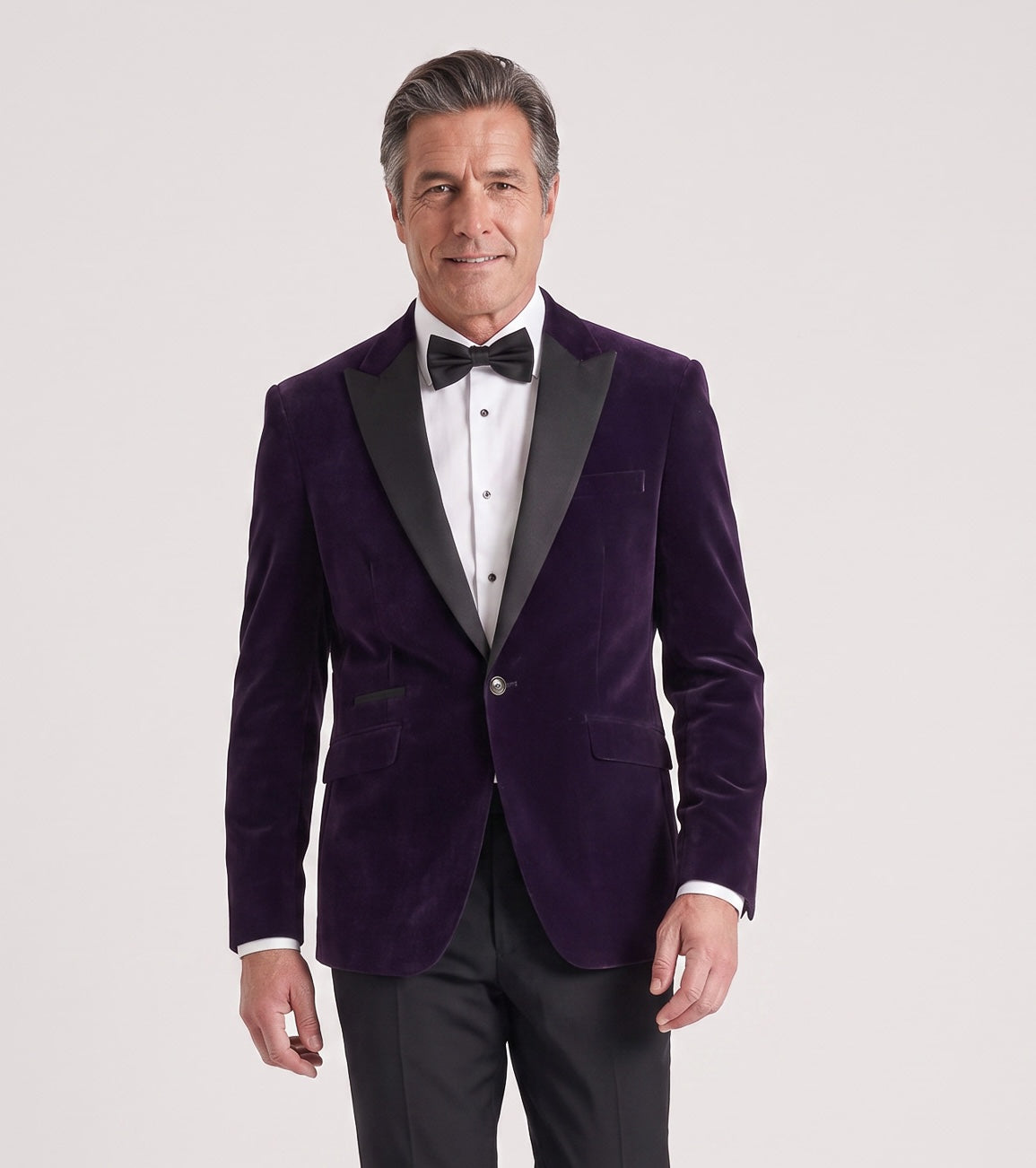 A man modeling a dark purple velvet tuxedo jacket with black satin lapels, paired with a white dress shirt, black bow tie, and black trousers, against a light gray studio background.
