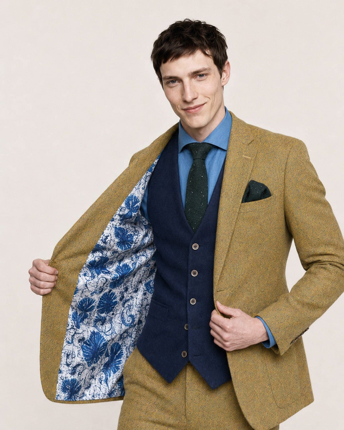 A man modeling a two-piece suit with a textured mustard-yellow tweed blazer and trousers, a matching waistcoat, a blue shirt, a dark green tie, and a blue-and-white patterned lining visible inside the open blazer.